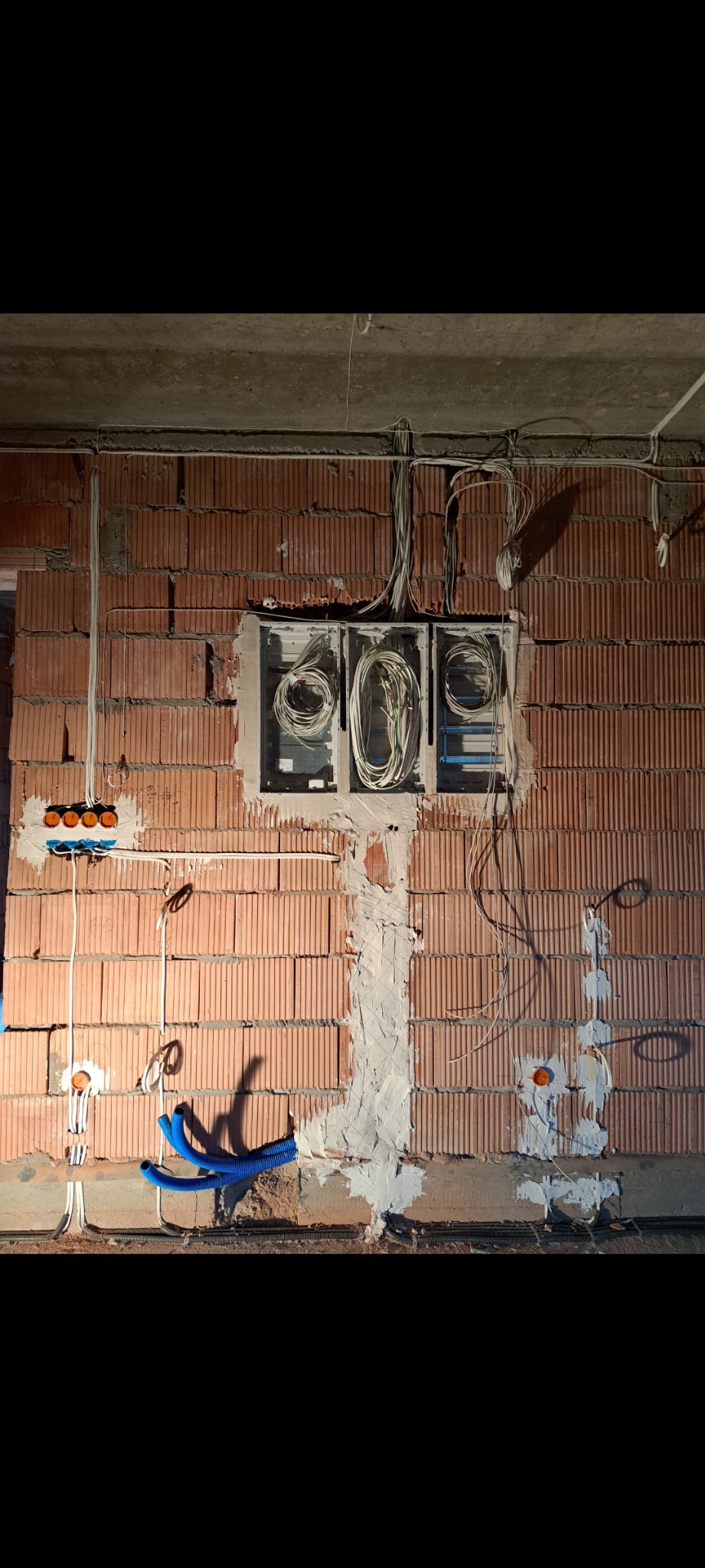 Instalacja elektryczna w trakcie montażu na ścianie z cegły. Widoczne kable w puszkach, rurki instalacyjne i fragment sufitu. Prace wewnątrz budynku.