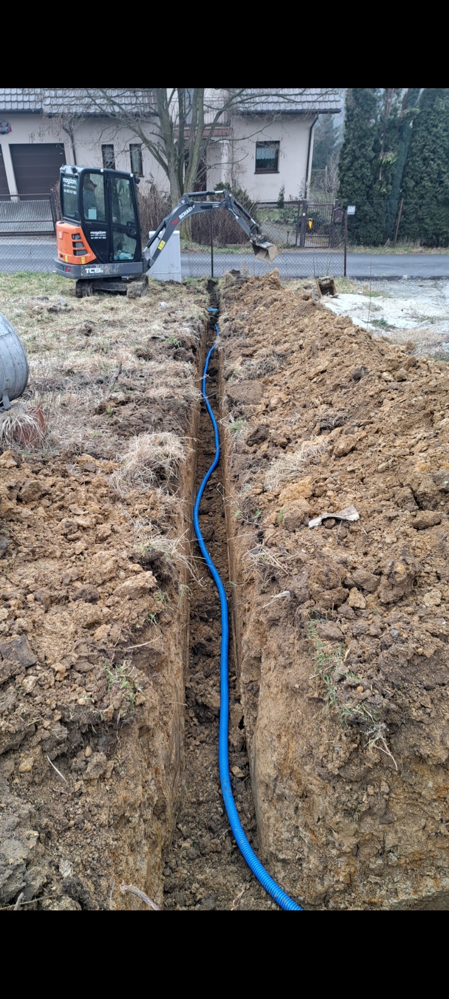 Niebieski przewód w wykopie ziemnym, widoczny na tle minikoparki i zabudowań. Instalacja podziemna w trakcie realizacji.
