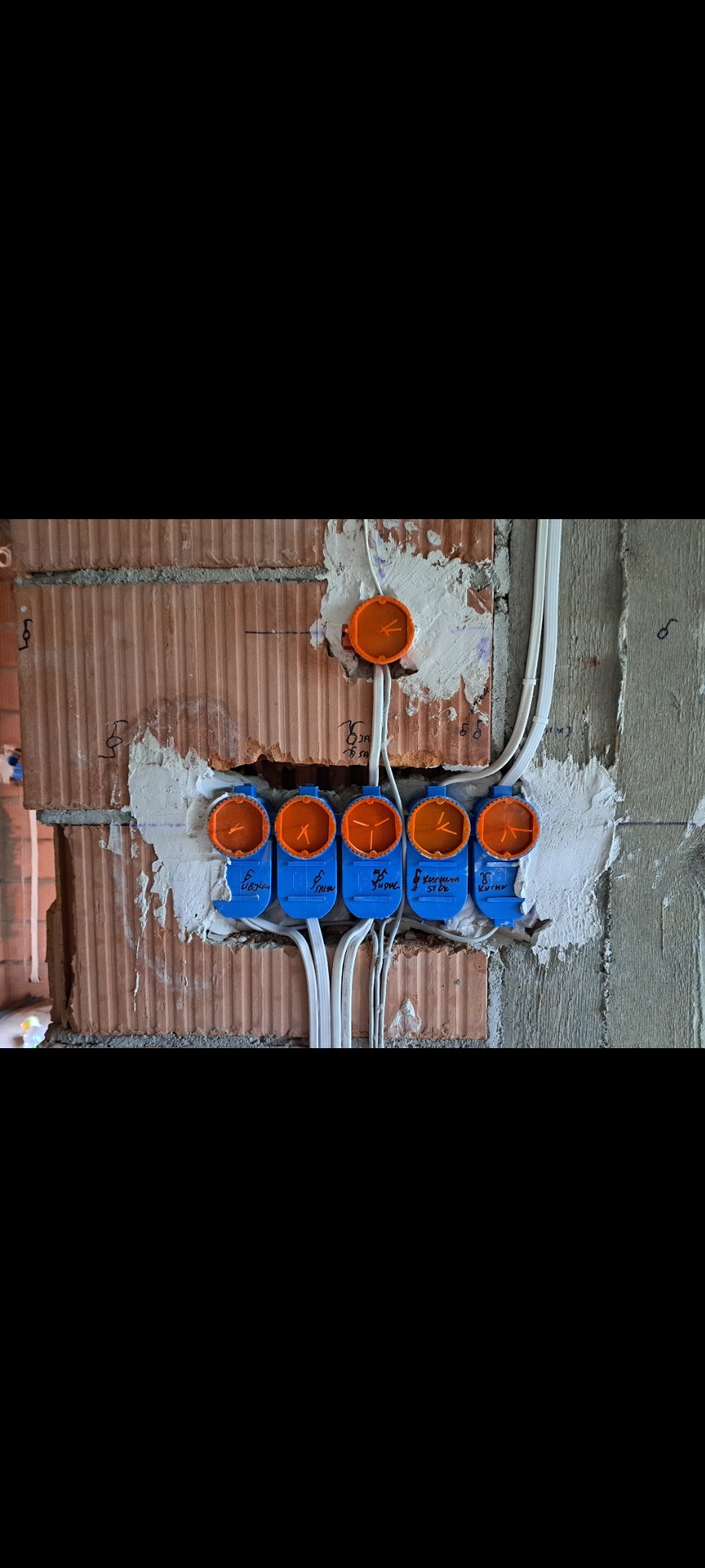 Instalacja elektryczna w trakcie montażu: widoczne puszki instalacyjne w kolorze pomarańczowym i niebieskim, przewody oraz surowa ściana z cegły.