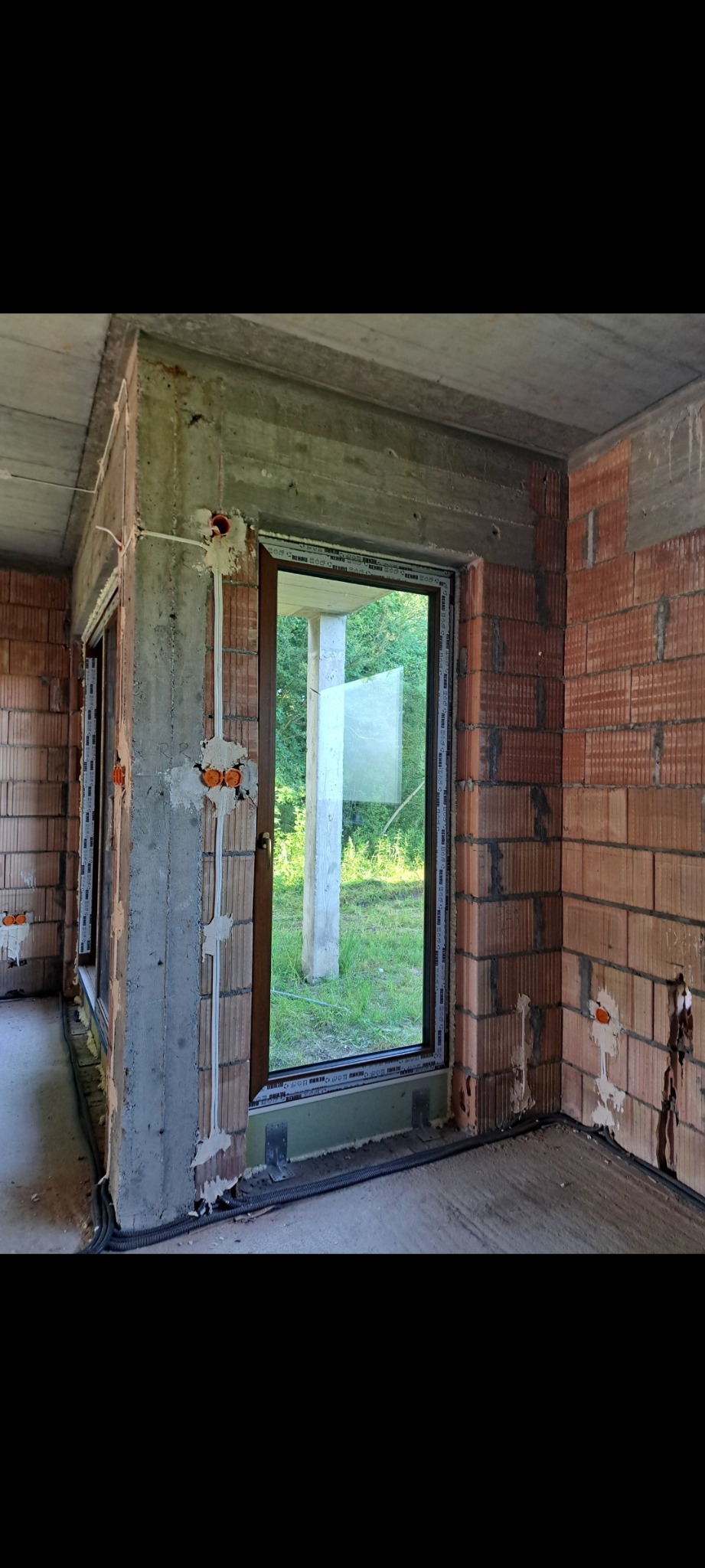 Surowy stan wnętrza z widoczną instalacją elektryczną: białe rurki z kablami prowadzone wzdłuż ściany, pomarańczowe puszki pod gniazdka, ściany z czerwonej cegły.