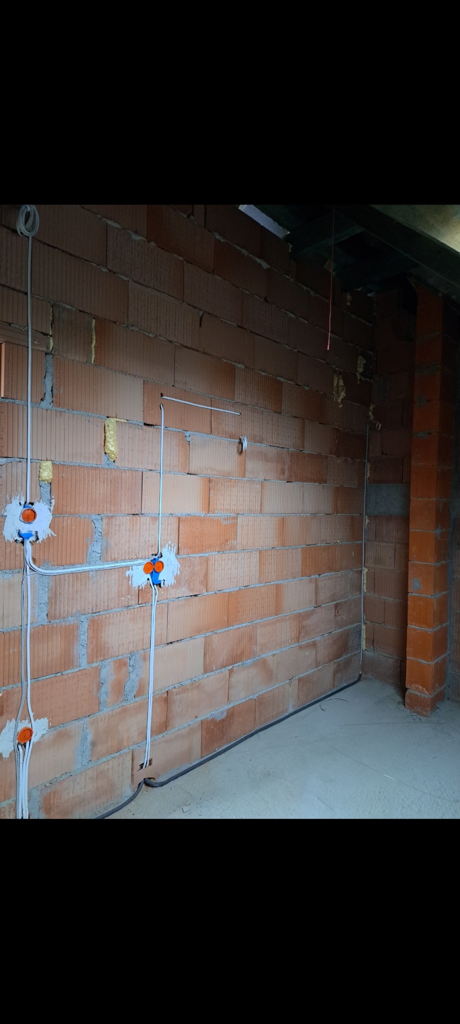 Ściana z cegły z zamontowanymi puszkami elektrycznymi i przewodami w trakcie instalacji. Widoczne przewody prowadzone po ścianie i pod podłogą.