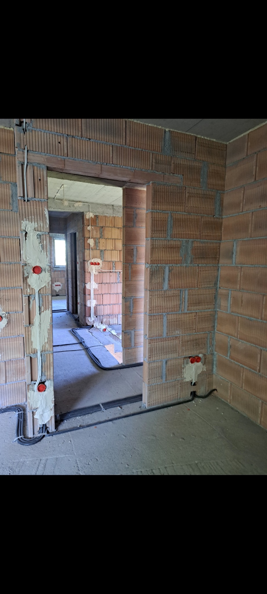 Wnętrze w trakcie prac: widoczne kable elektryczne prowadzone po podłodze i w ścianach z czerwonej cegły, gniazda elektryczne w puszkach instalacyjnych.