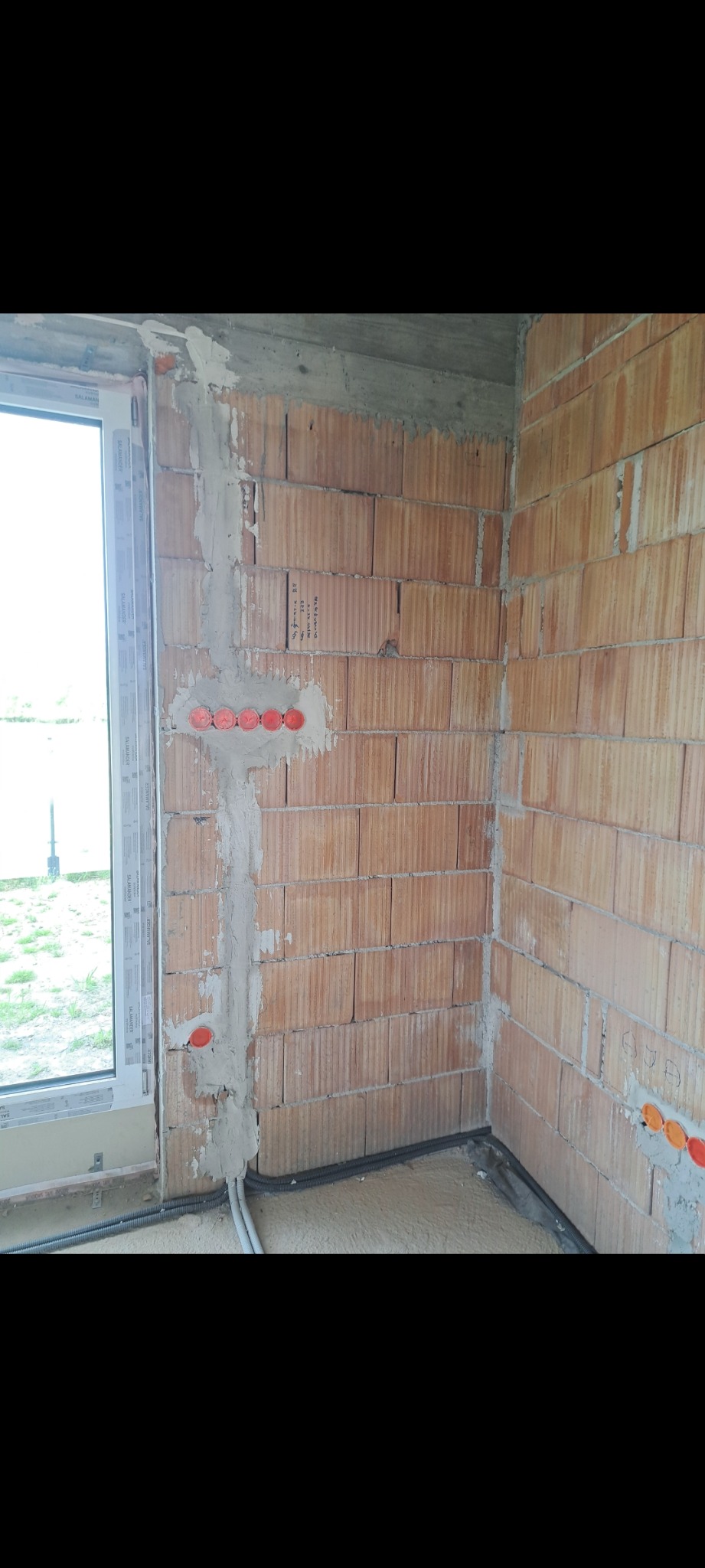 Wewnętrzna instalacja elektryczna w trakcie montażu w surowym budynku. Widoczne kable w peszlach, puszki podtynkowe zalane gipsem i fragment okna.