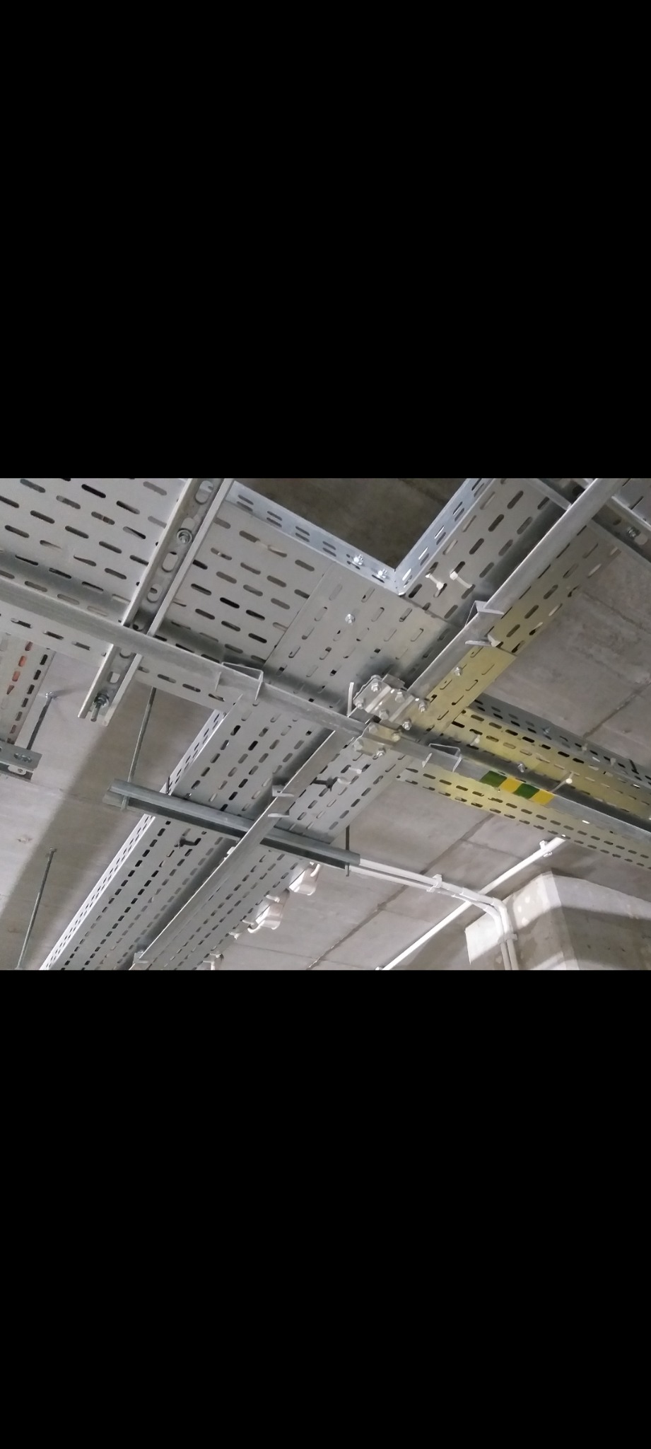 Metalowe korytka kablowe, perforowane, zamocowane pod sufitem. Widoczne białe rury instalacyjne. Szare, betonowe tło sufitu. Kąt ujęcia od dołu.
