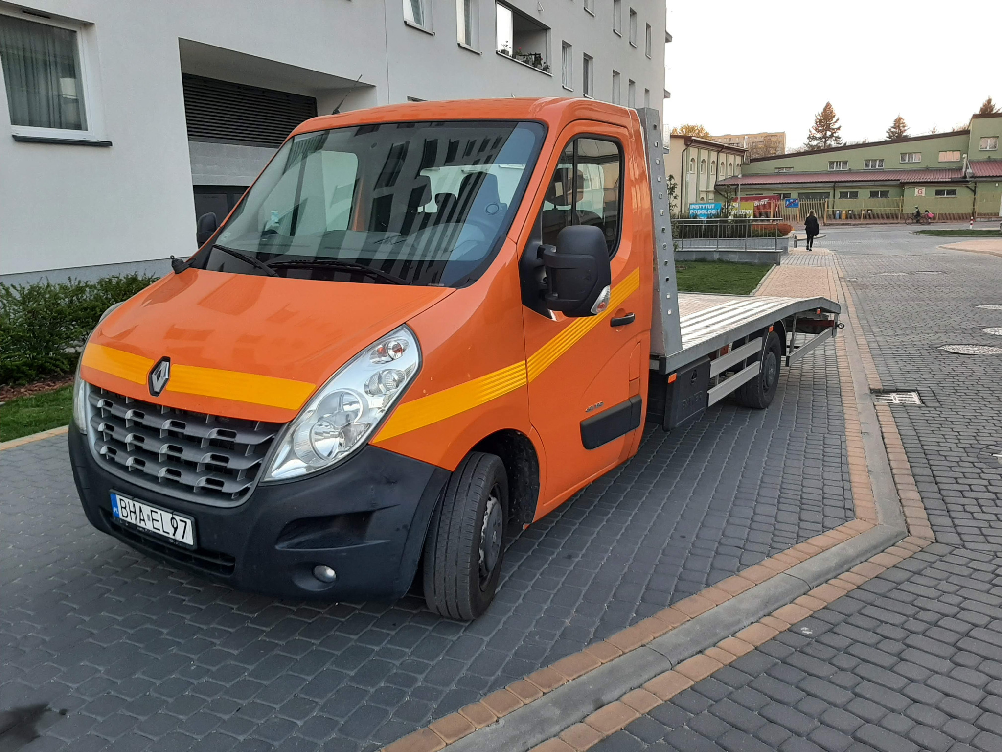 Pomarańczowa laweta Renault Master z żółtym pasem na boku, zaparkowana na kostce brukowej przed budynkiem.
