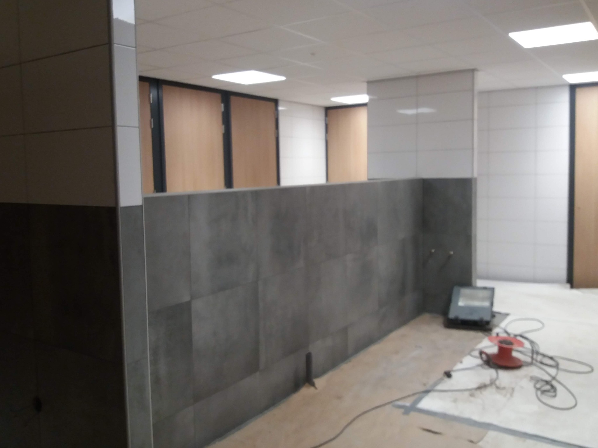 Szare płytki na ścianie w trakcie montażu, widoczne narzędzia i kable na podłodze, fragmenty białych płytek na górze i drzwi w tle.