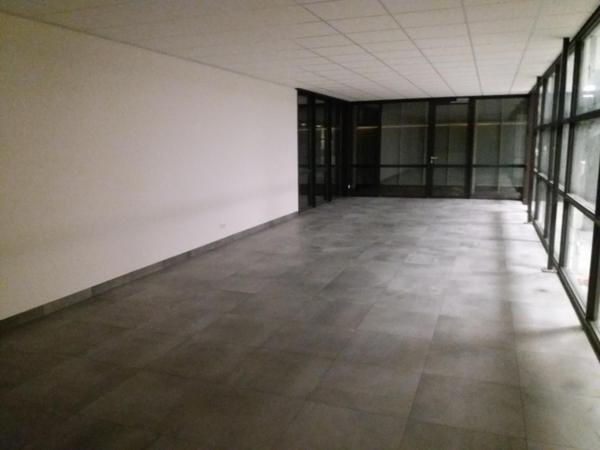 Pusta przestrzeń biurowa z ciemnoszarą podłogą z płytek, białą ścianą i przeszklonymi ścianami z czarnymi ramami.