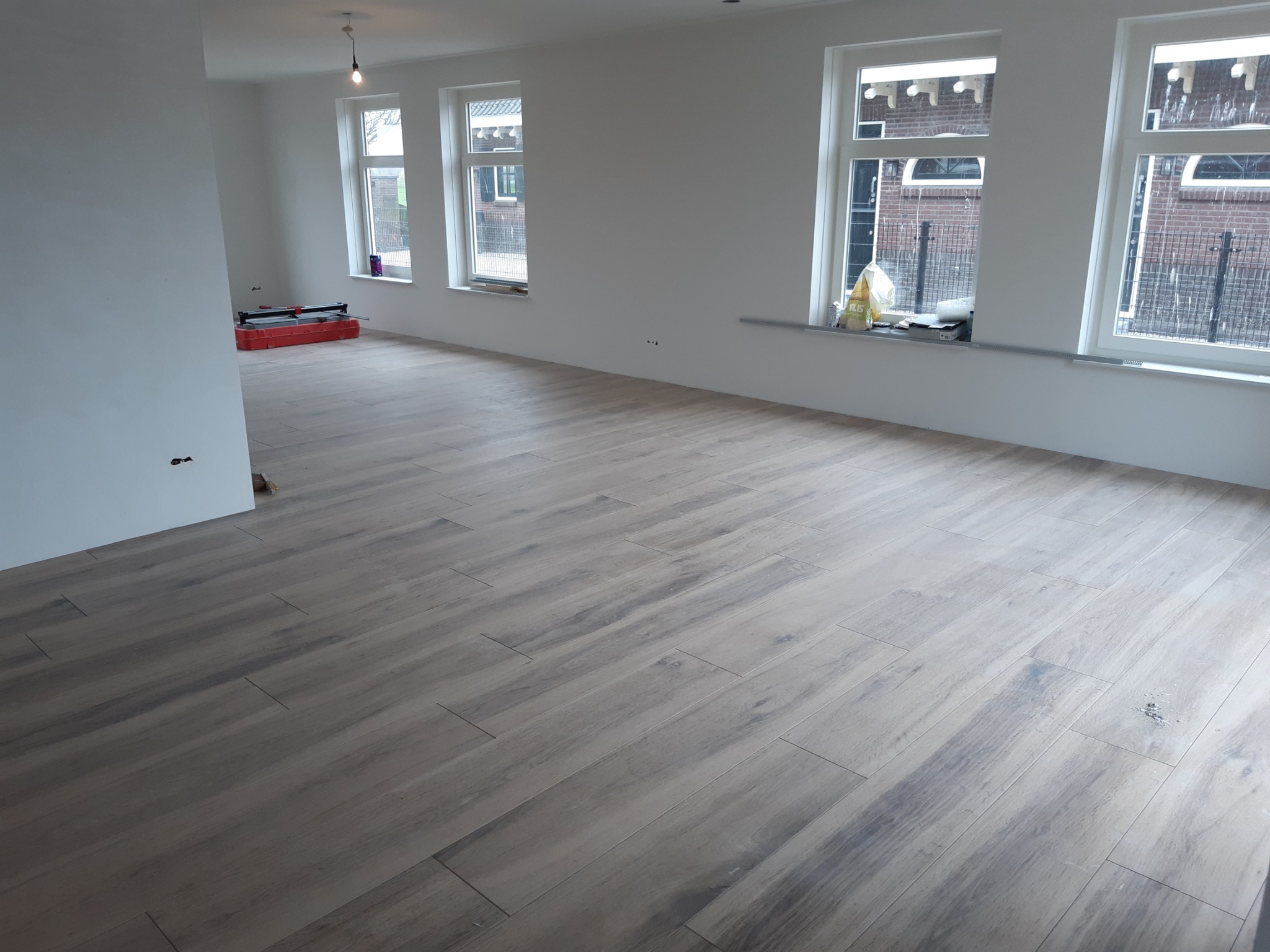 Ułożona podłoga z płytek imitujących drewno w jasnym, nowo wykończonym wnętrzu. W tle widoczny czerwony sprzęt do cięcia płytek i okna z widokiem na budynki.