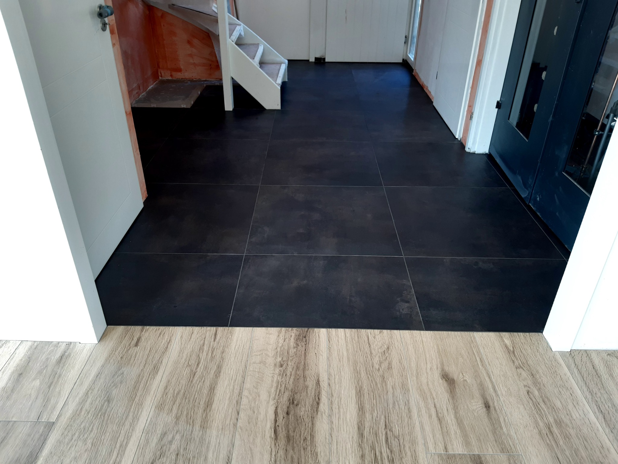 Ułożona, ciemna, matowa podłoga z płytek w korytarzu, z widocznym fragmentem schodów i różnicą poziomów z panelami podłogowymi w innym pomieszczeniu.