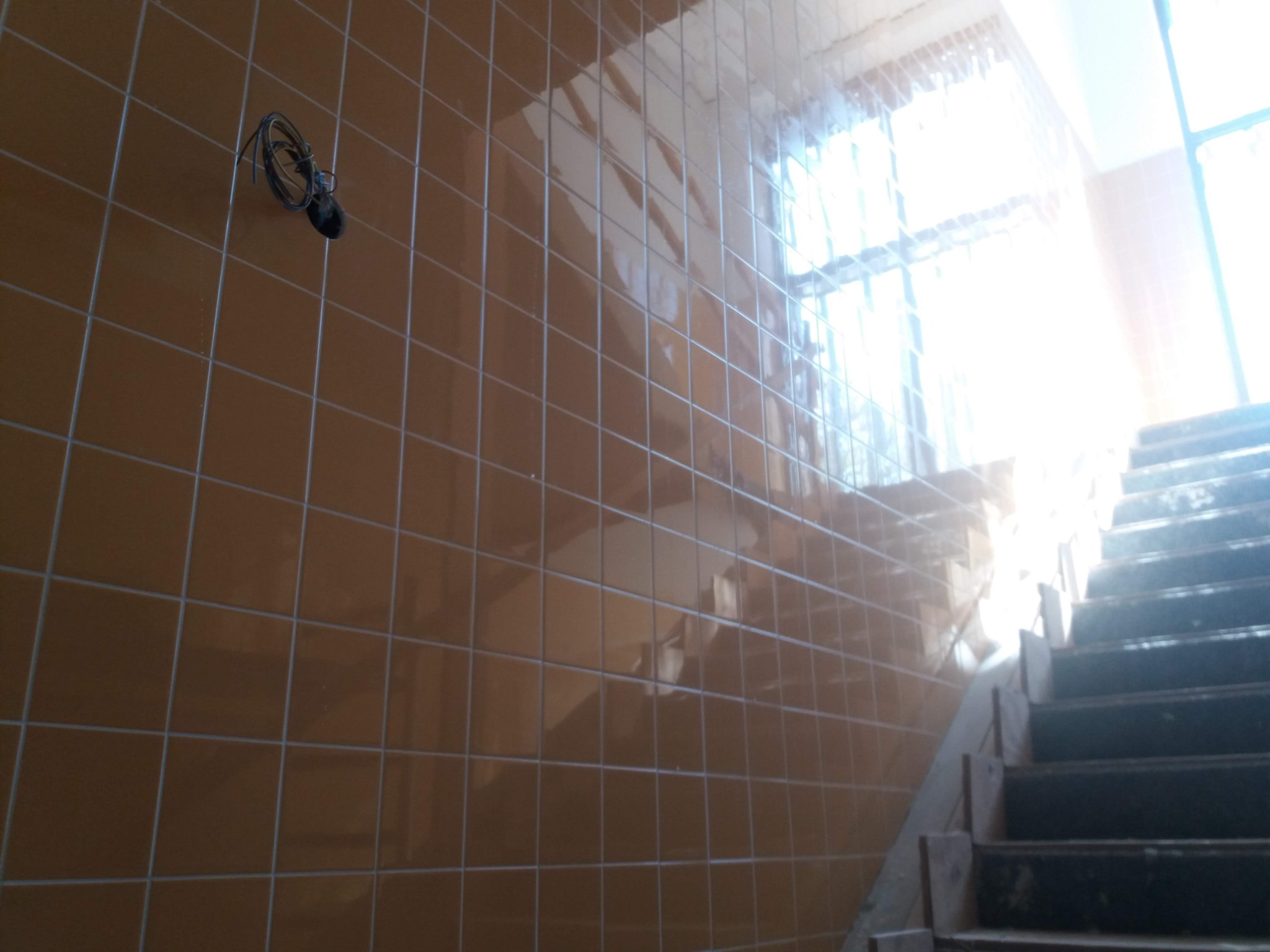 Klatka schodowa wyłożona błyszczącymi, brązowymi płytkami ceramicznymi z widocznymi przewodami elektrycznymi na ścianie.