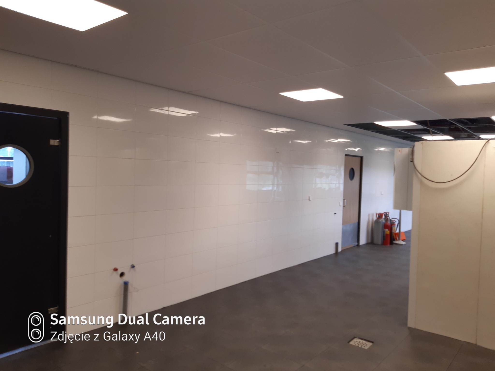 Wnętrze z białymi płytkami na ścianach, szarą podłogą i sufitem z oświetleniem panelowym, widoczne przyłącza wodne i kanalizacyjne oraz drzwi po lewej stronie.