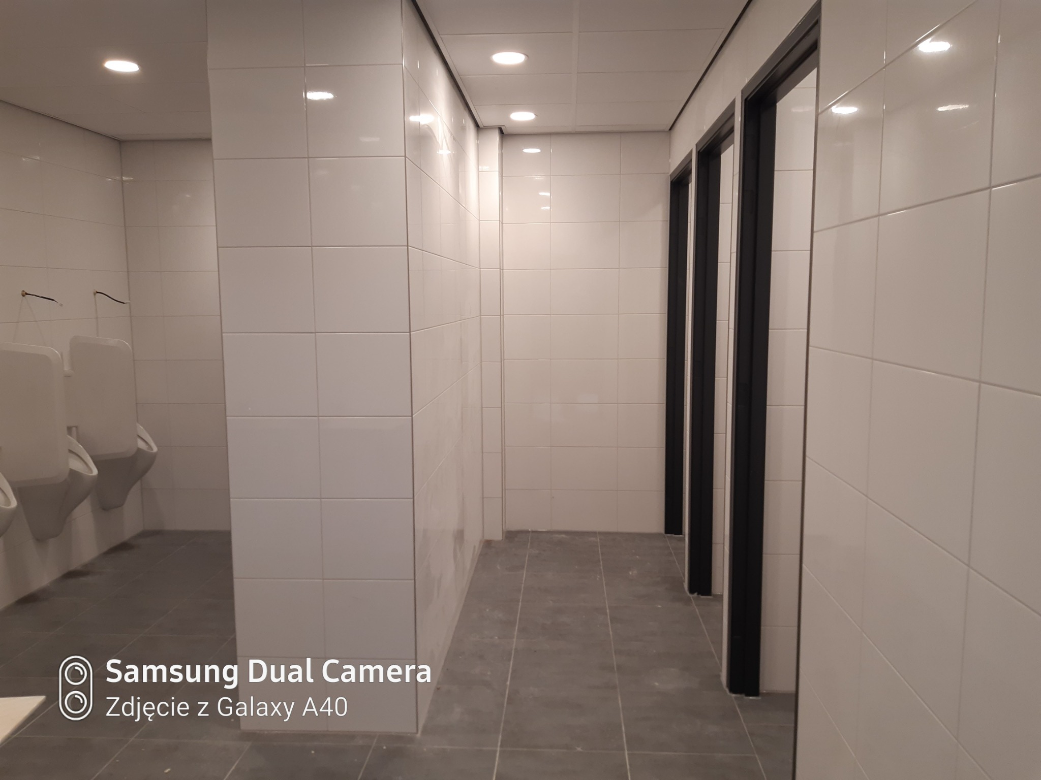 Wykończona łazienka publiczna z białymi płytkami na ścianach i szarymi na podłodze, widoczne pisuary i czarne ramy drzwi do kabin.