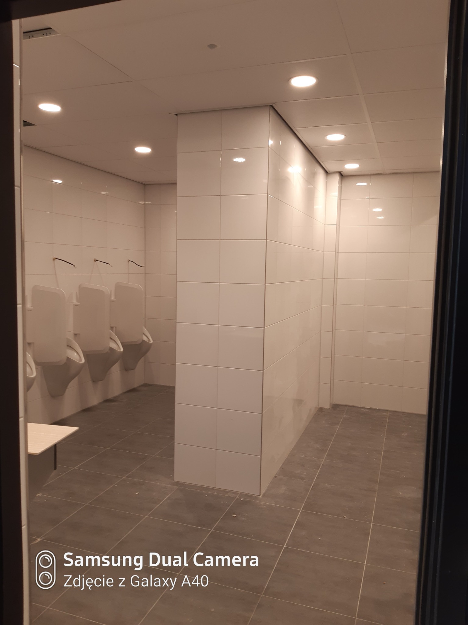 Wykończona toaleta publiczna z białymi, kwadratowymi płytkami na ścianach i szarymi płytkami podłogowymi, widoczne pisuary.