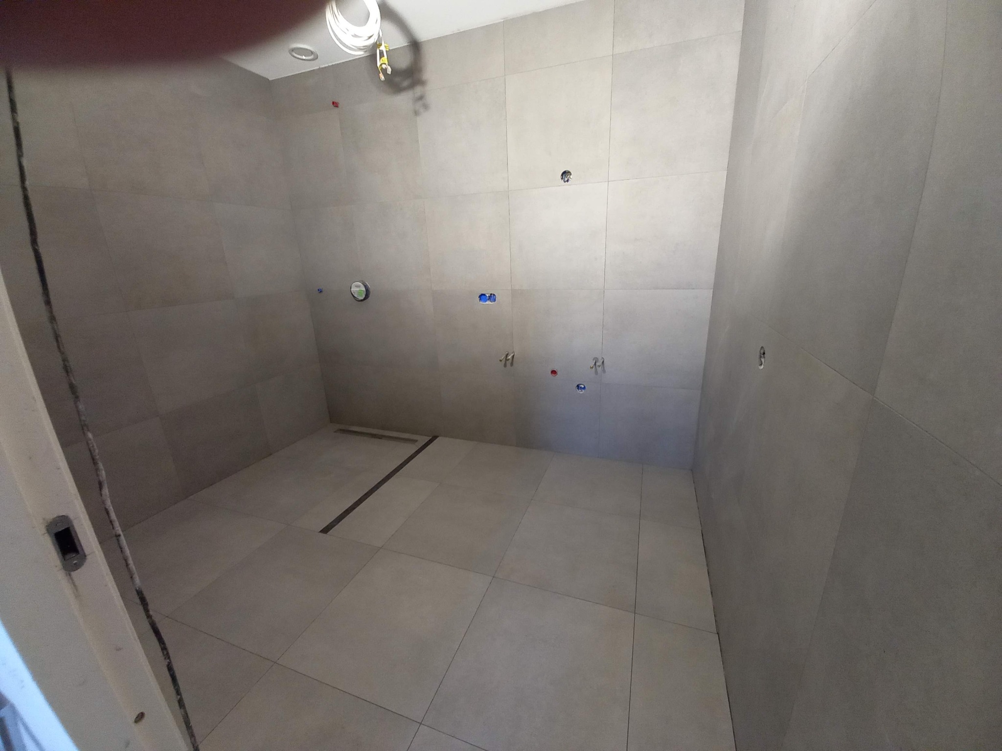 Łazienka w trakcie wykańczania: szare, matowe płytki na ścianach i podłodze, widoczne punkty instalacji, odpływ liniowy w podłodze.