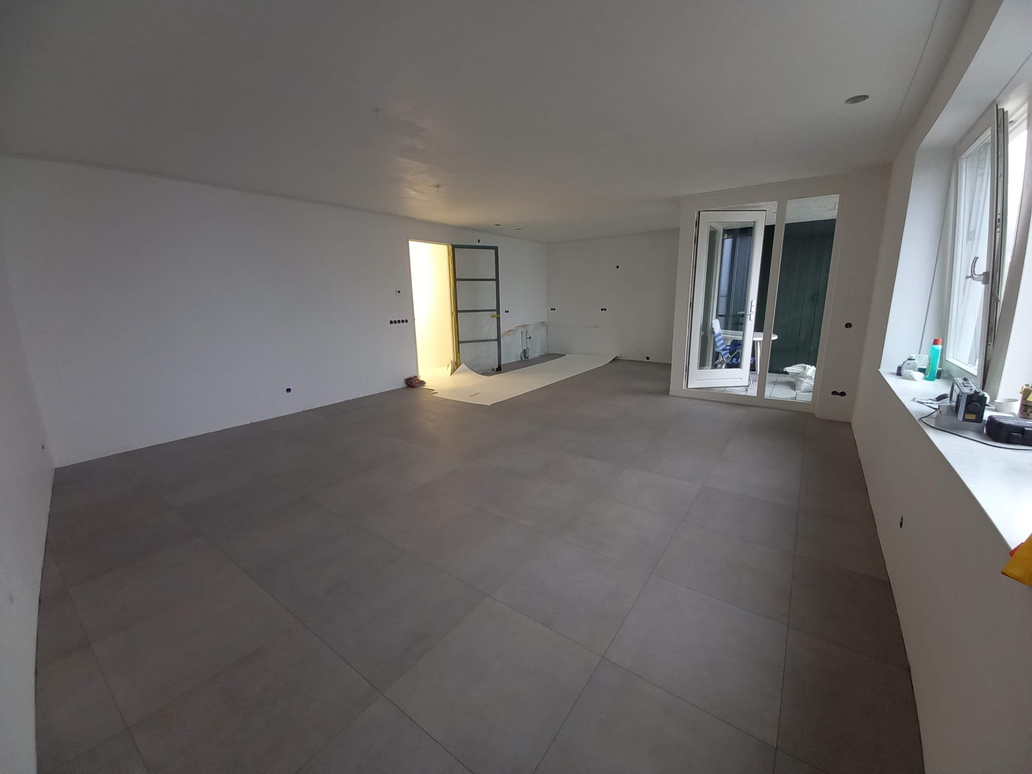 Układanie szarych płytek podłogowych w dużym, jasnym pomieszczeniu z białymi ścianami, widoczne przygotowane podłoże w przejściu do kolejnego pokoju i narzędzia na parapecie.