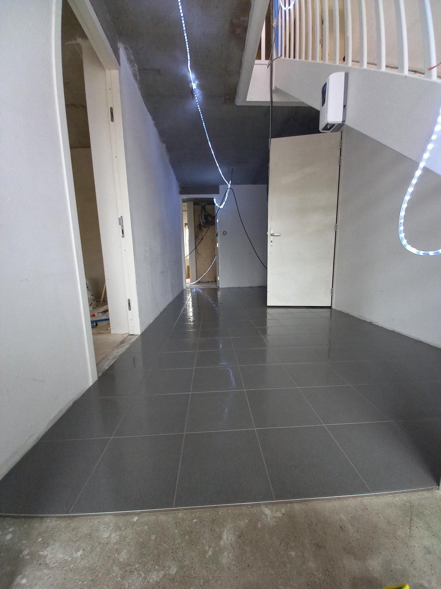 Ułożona, ciemnoszara podłoga z płytek w korytarzu, widok na próg z metalową listwą, w tle niedokończone ściany i instalacje.