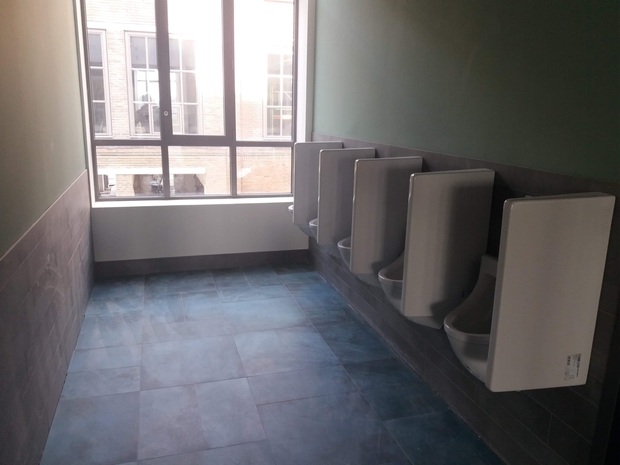 Rząd białych pisuarów w publicznej toalecie z szarą glazurą na ścianach i podłodze, duże okno z widokiem na budynek z cegły.