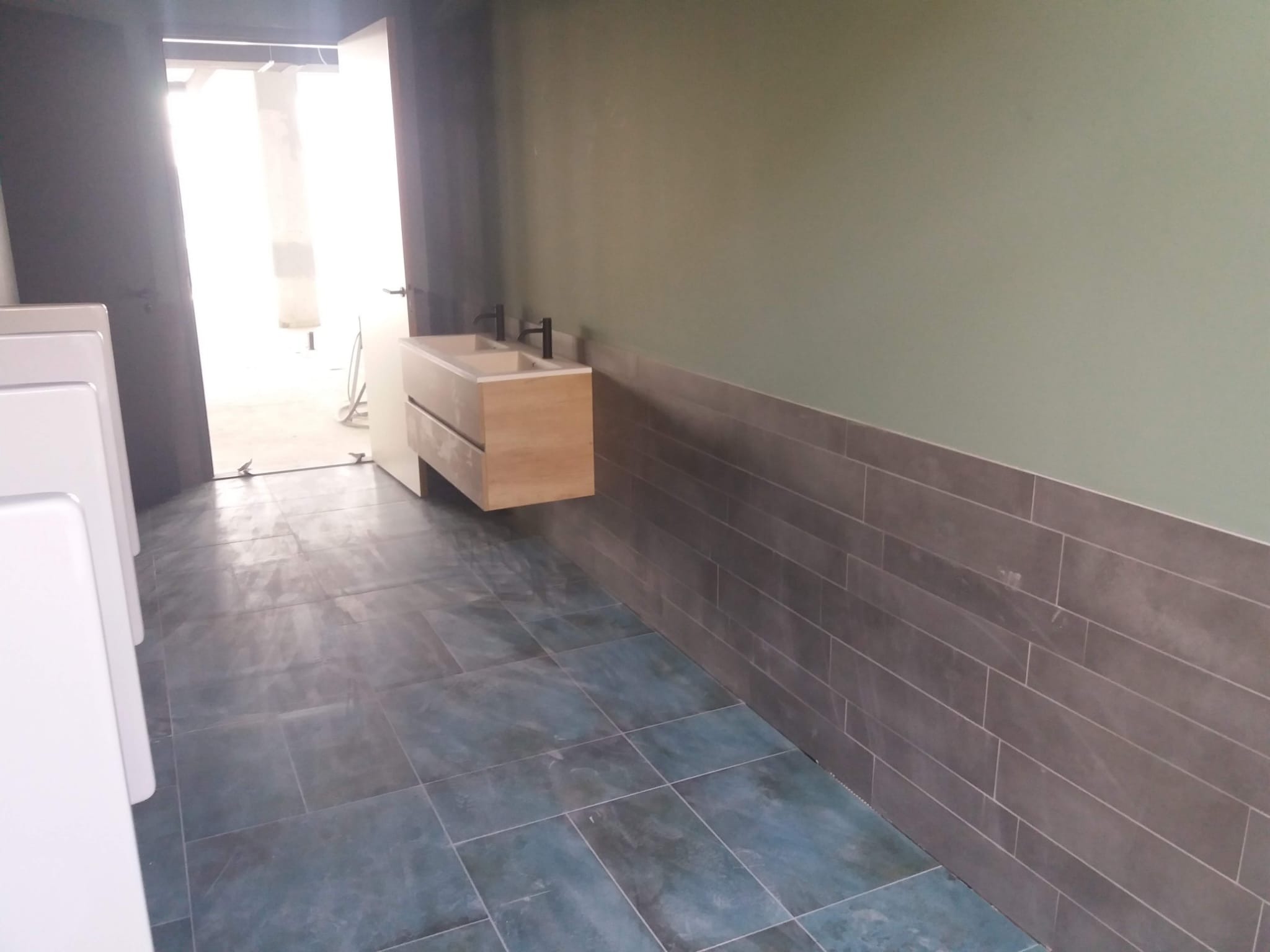 Nowoczesna łazienka z szaro-zielonymi ścianami i podłogą z płytek w odcieniach niebieskiego, z wiszącą szafką z umywalkami i czarną armaturą.