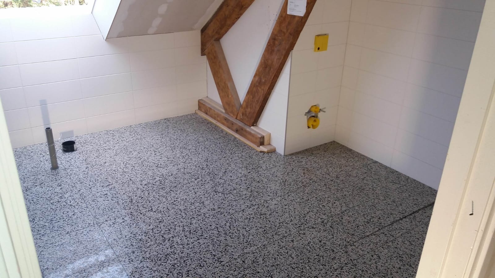 Wykończona łazienka na poddaszu z szarą, nakrapianą posadzką żywiczną, białe płytki na ścianach, widoczne drewniane elementy konstrukcyjne, rura odpływowa i żółte przyłącza wodne.
