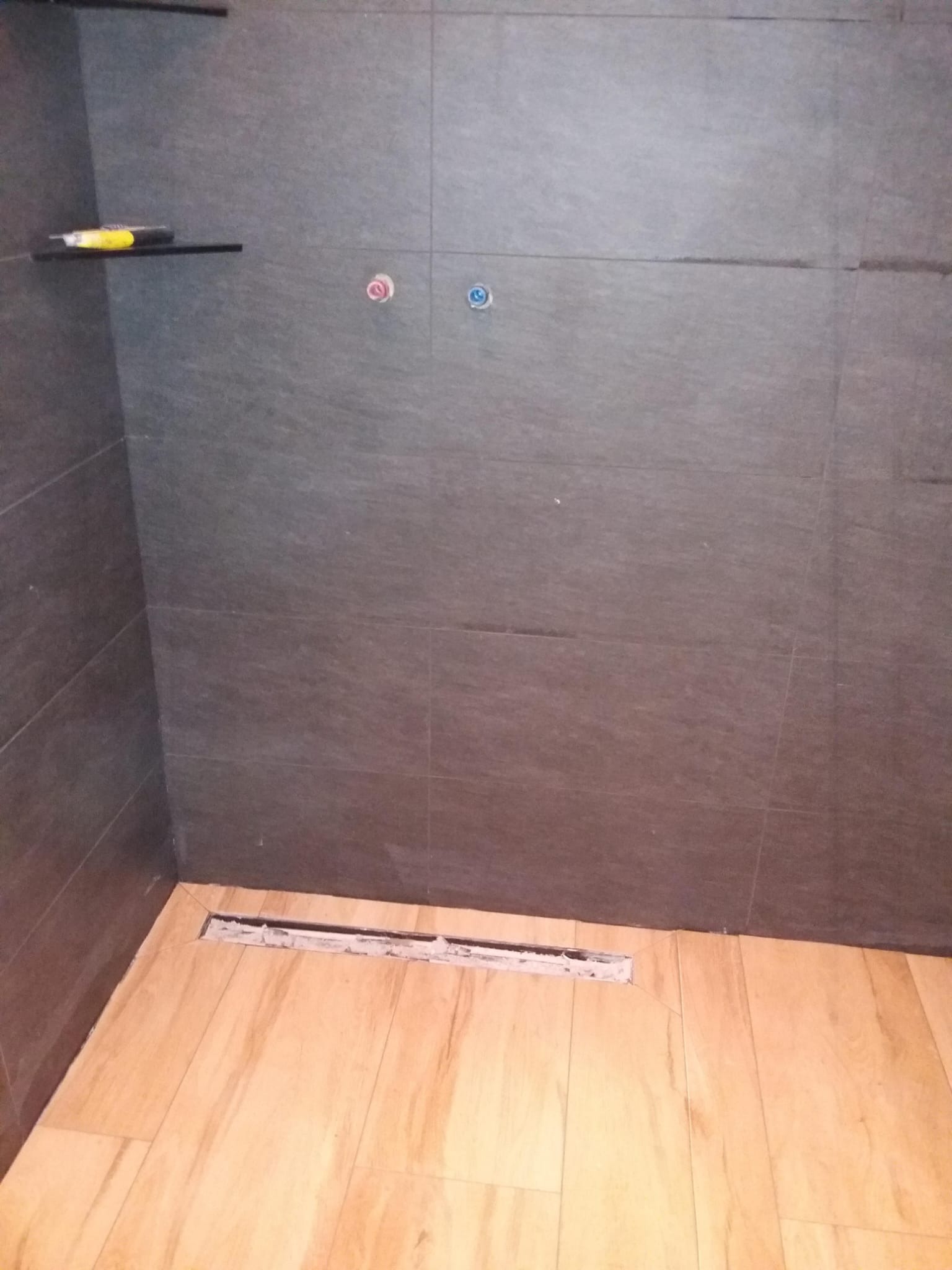 Ułożona glazura w kabinie prysznicowej z odpływem liniowym, widoczne przyłącza wodne i półka z narzędziem.