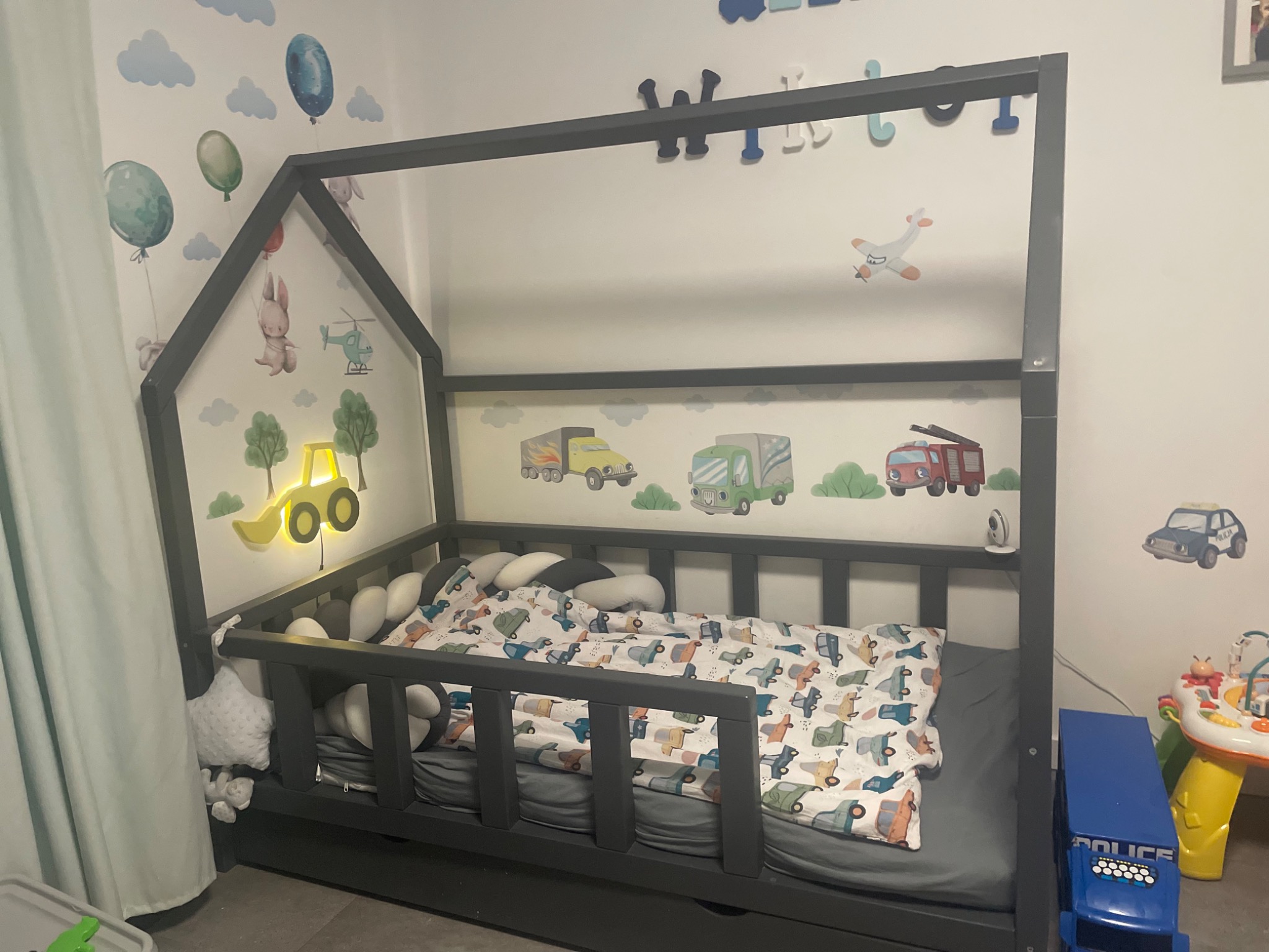 Szare łóżko domek dla dziecka z barierkami, pościel w pojazdy, poduszki wałki, naklejki na ścianie: balony, zwierzęta, pojazdy, świecąca lampka w kształcie koparki.