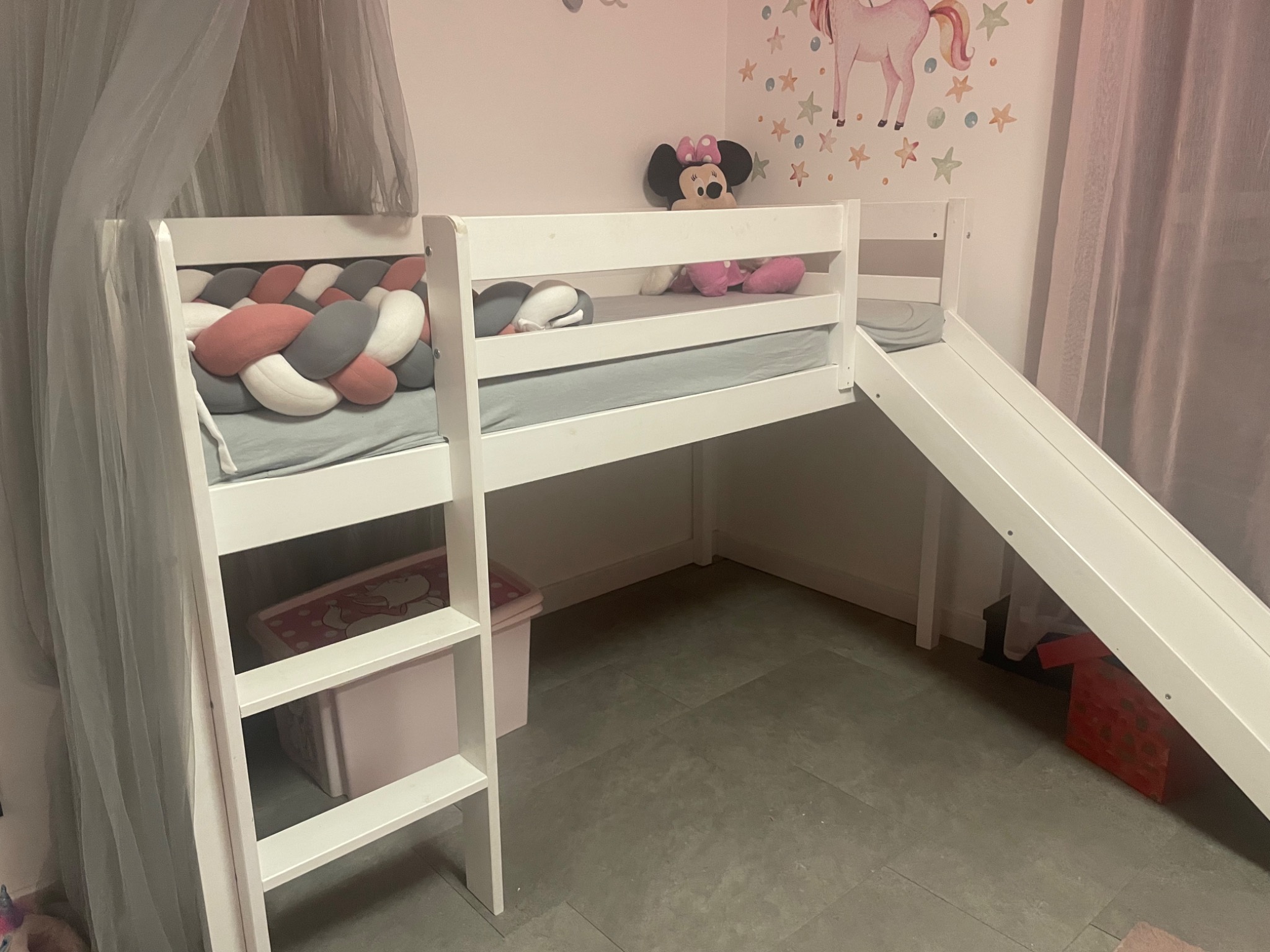 Białe łóżko piętrowe dla dziecka z drabinką, zjeżdżalnią i baldachimem, ozdobione pluszową poduszką w warkocz i naklejkami na ścianie z jednorożcem i gwiazdkami.