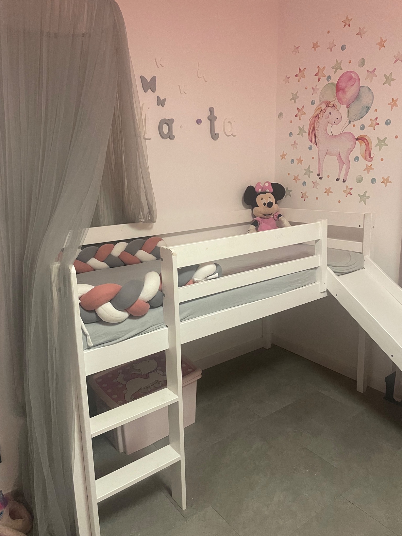 Białe łóżko dziecięce na podeście z drabinką i zjeżdżalnią, ozdobione plecionym ochraniaczem i pluszową zabawką Myszki Minnie, na ścianie naklejki z jednorożcem i gwiazdkami.