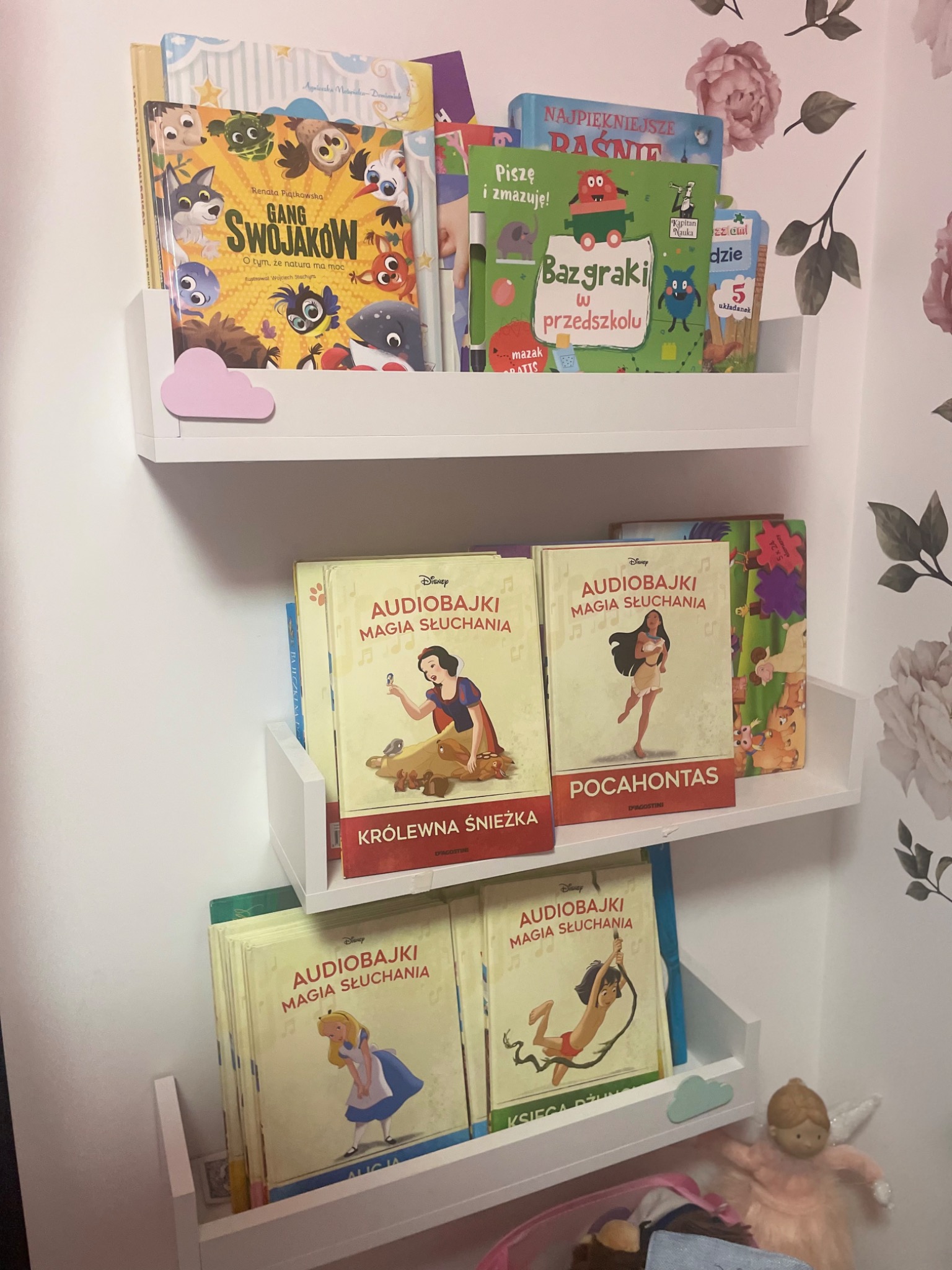 Trzy białe półki na książki w pokoju dziecięcym, z książkami dla dzieci, w tym 'Gang Swojaków', 'Bazgraki w przedszkolu' i seria 'Audiobajki Magia Słuchania' z Królewną Śnieżką, Pocahontas, Alicją...