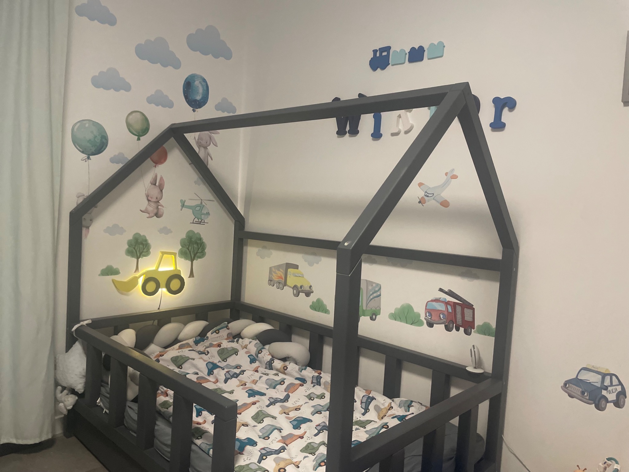 Szare łóżko domek dla dziecka z pościelą w samochody, na ścianie naklejki z balonami, chmurami, zwierzętami, pojazdami i podświetlana lampka w kształcie koparki.