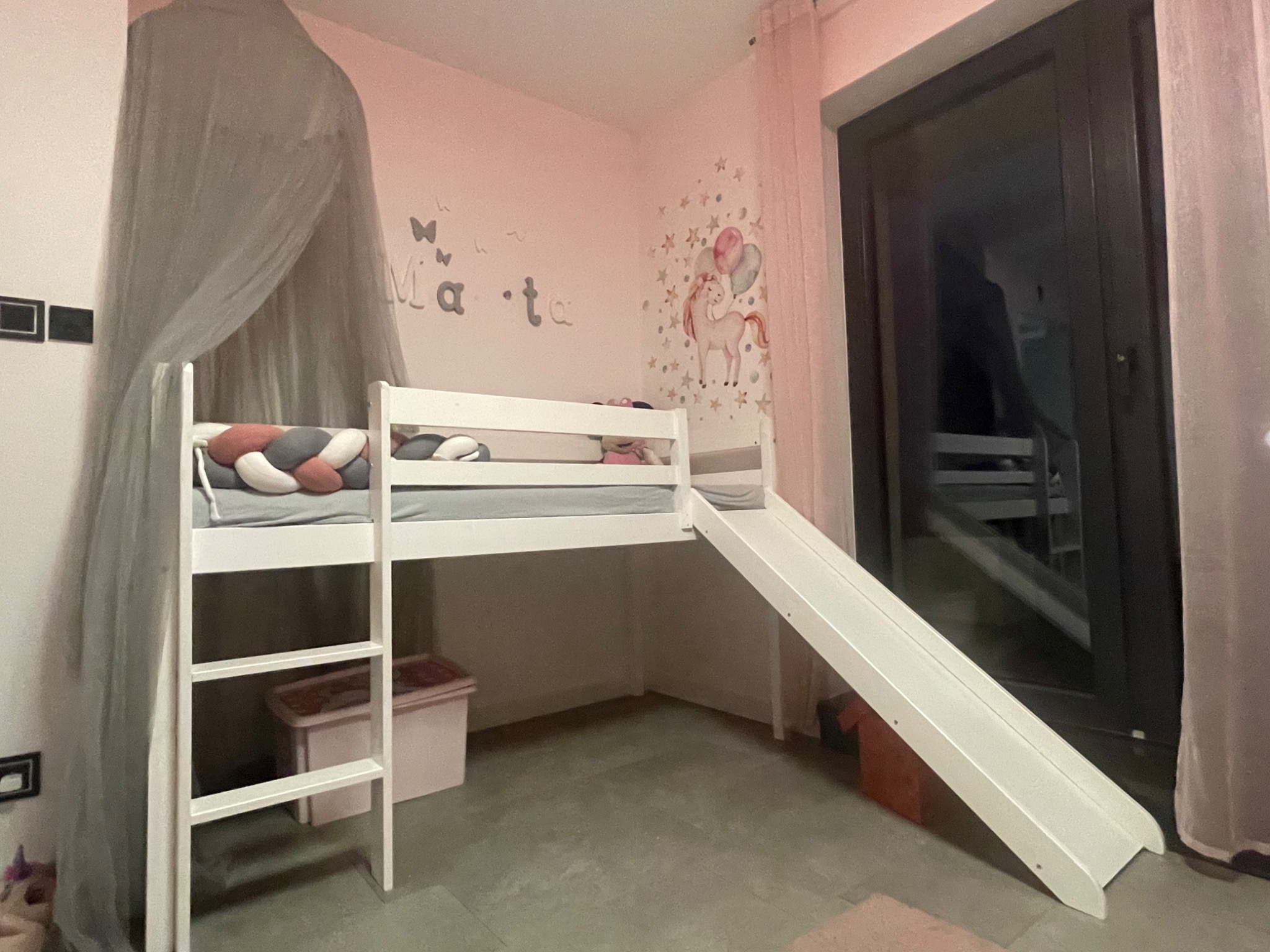 Białe łóżko piętrowe z drabinką i zjeżdżalnią w pokoju dziecięcym z różowymi ścianami i naklejkami z jednorożcem i imieniem 'Marta'.