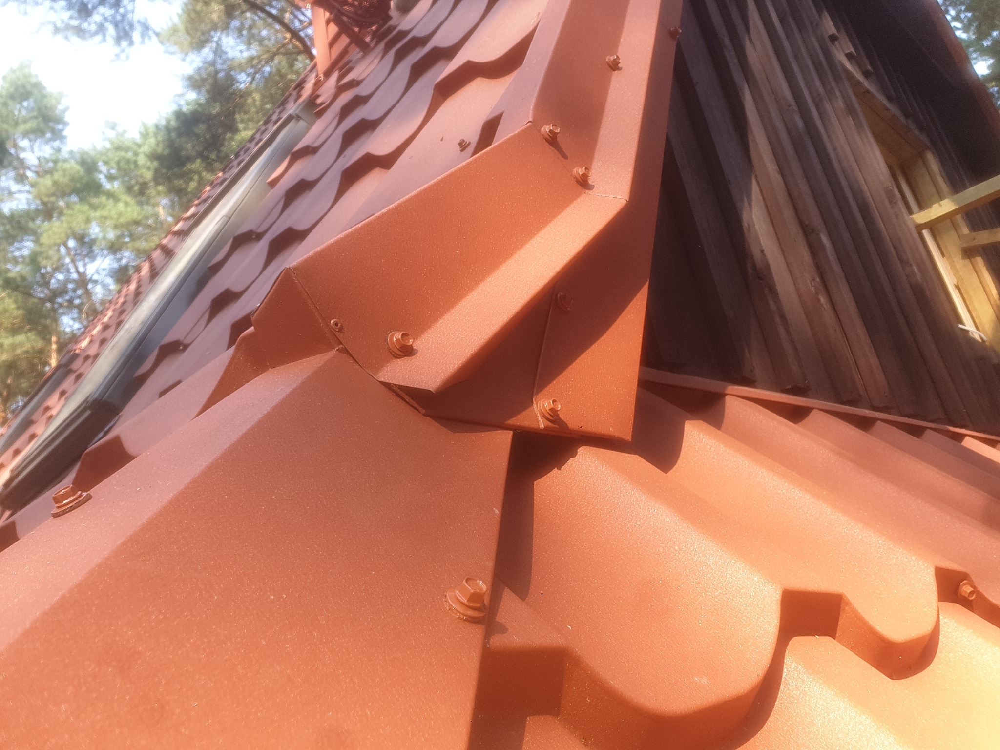 Szczegółowe ujęcie pomarańczowo-brązowego dachu z blachy trapezowej, z widocznymi mocowaniami śrubowymi i obróbkami blacharskimi przy oknie dachowym i ścianie budynku.