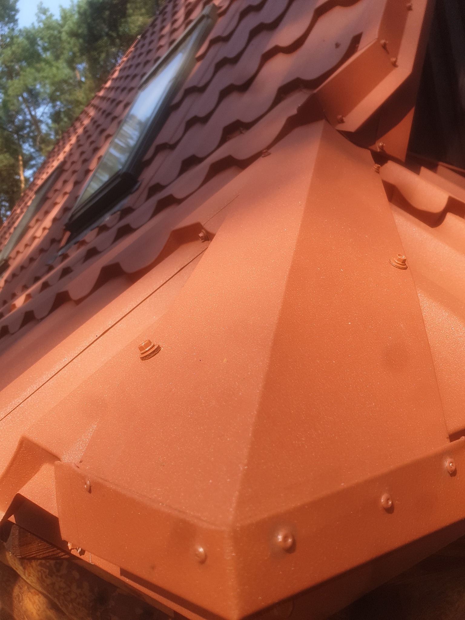 Pomarańczowo-czerwone, metalowe obróbki blacharskie na dachu z dachówką, widoczne okno dachowe i drzewa w tle.