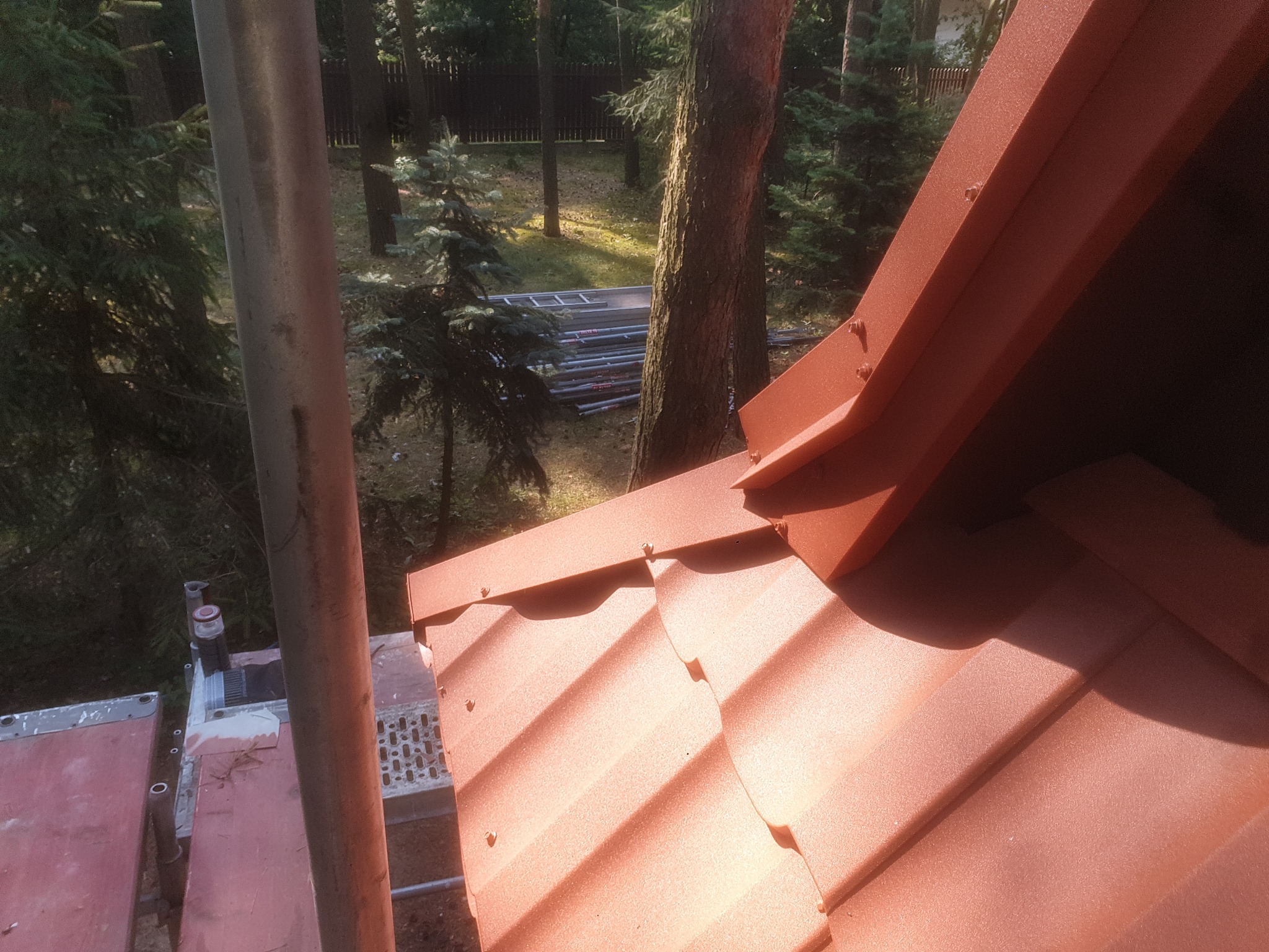 Widok z góry na fragment dachu z blachy w kolorze ceglastym, widoczne łączenia i mocowania, w tle drzewa iglaste i drabina oparta o elewację.