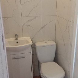 Jarmarhome - Mała łazienka z białymi marmurkowymi płytkami ściennymi, brązowymi płytkami podłogowymi, białą szafką z umywalką i toaletą.