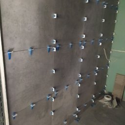 Jarmarhome - Świeżo ułożone, szare płytki ścienne z niebiesko-białymi klipsami poziomującymi i cienkimi żyłkami, widoczne resztki materiałów budowlanych na podłodze.