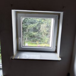 Jarmarhome - Okno dachowe po montażu, zabezpieczone taśmą malarską, z widokiem na zieleń ogrodu.