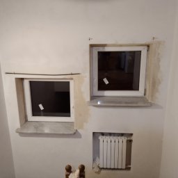 Jarmarhome - Dwa okna PCV z marmurowymi parapetami osadzone w ścianie, widoczne ślady po szpachlowaniu wokół ram, pod jednym z okien grzejnik.