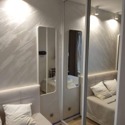Jarmarhome - Sypialnia z łóżkiem, lustrem na ścianie i szafą przesuwną z lustrzanymi drzwiami, wzorzysta ściana w odcieniach beżu, widoczny fragment podłogi i narzędzie malarskie.