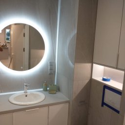 Jarmarhome - Nowoczesna łazienka z podświetlanym lustrem, umywalką na szafce, szarą glazurą imitującą marmur i zabudowaną toaletą z niebieską taśmą zabezpieczającą.