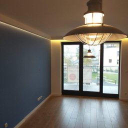 Jarmarhome - Pusta przestrzeń po remoncie: ściana pomalowana na niebiesko z listwą LED pod sufitem, parkiet ułożony na podłodze, duże okno balkonowe i designerska lampa w stylu industrialnym.