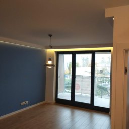 Jarmarhome - Wnętrze pokoju po remoncie: ściana pomalowana na niebiesko, jasna drewniana podłoga, duże okno balkonowe z czarną ramą, nowoczesna lampa wisząca, listwa LED pod sufitem.