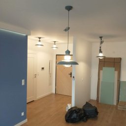 Jarmarhome - Wnętrze mieszkania po malowaniu ścian na kolor niebieski, z zamontowanym oświetleniem sufitowym w stylu industrialnym, widoczne również panele podłogowe i worki na śmieci.