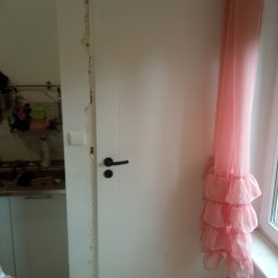 Jarmarhome - Białe drzwi wewnętrzne z czarną klamką, osadzone w ościeżnicy z widocznymi śladami pianki montażowej i szpachli, obok okna z różową firanką w falbany.