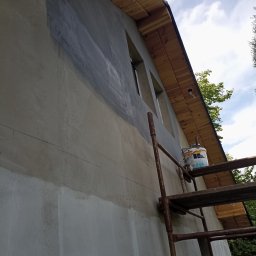 Jarmarhome - Ściana budynku w trakcie przygotowania do malowania, widoczne ślady szpachlowania i gruntowania, rusztowanie z wiadrem farby obok okien.