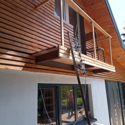 Jarmarhome - Dom z nową drewnianą elewacją i dobudowanym balkonem, widoczna antena TV oparta o ścianę, odbicie osoby w oknie.