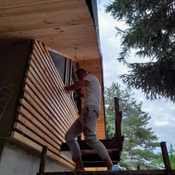 Jarmarhome - Pracownik na rusztowaniu mocuje drewniane listwy elewacyjne do ściany budynku, widoczne elementy konstrukcyjne domu i otaczająca zieleń.