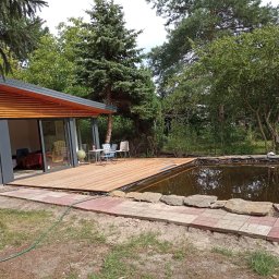 Jarmarhome - Nowoczesny domek letniskowy z drewnianym tarasem nad oczkiem wodnym, otoczony zielenią i brukowaną ścieżką