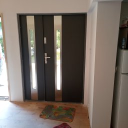 Jarmarhome - Nowe, grafitowe drzwi wejściowe z przeszkleniami, zamontowane w surowym stanie wykończeniowym, widoczne drewniane podłoże i dwa dywaniki.