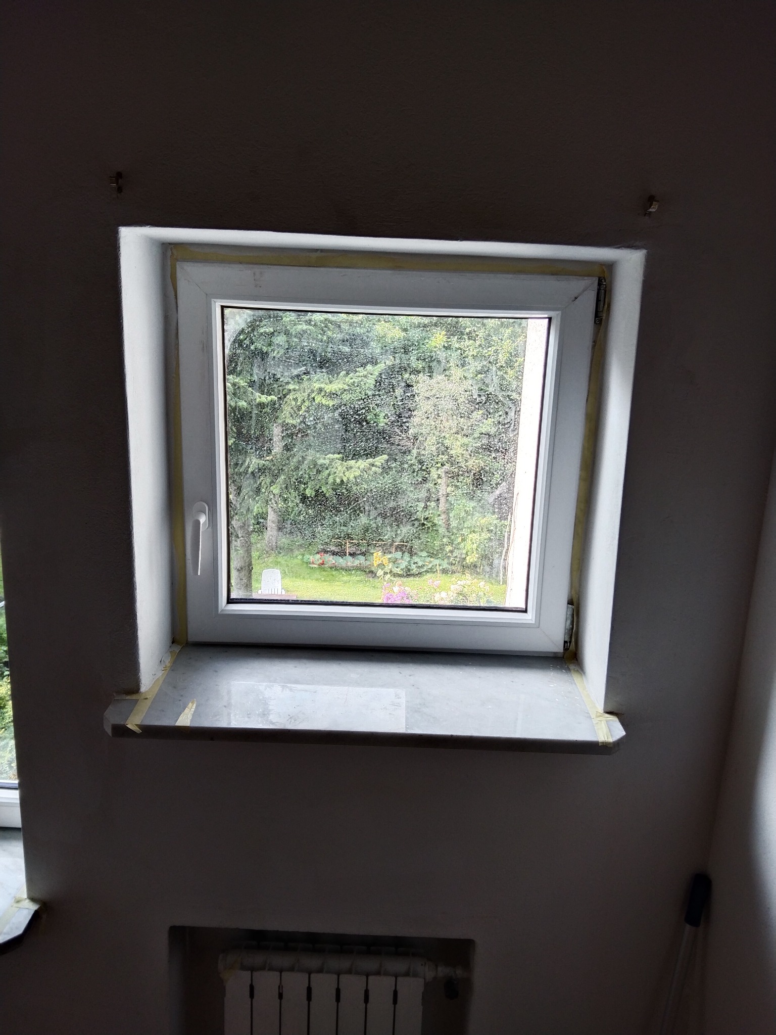 Okno dachowe po montażu, zabezpieczone taśmą malarską, z widokiem na zieleń ogrodu.