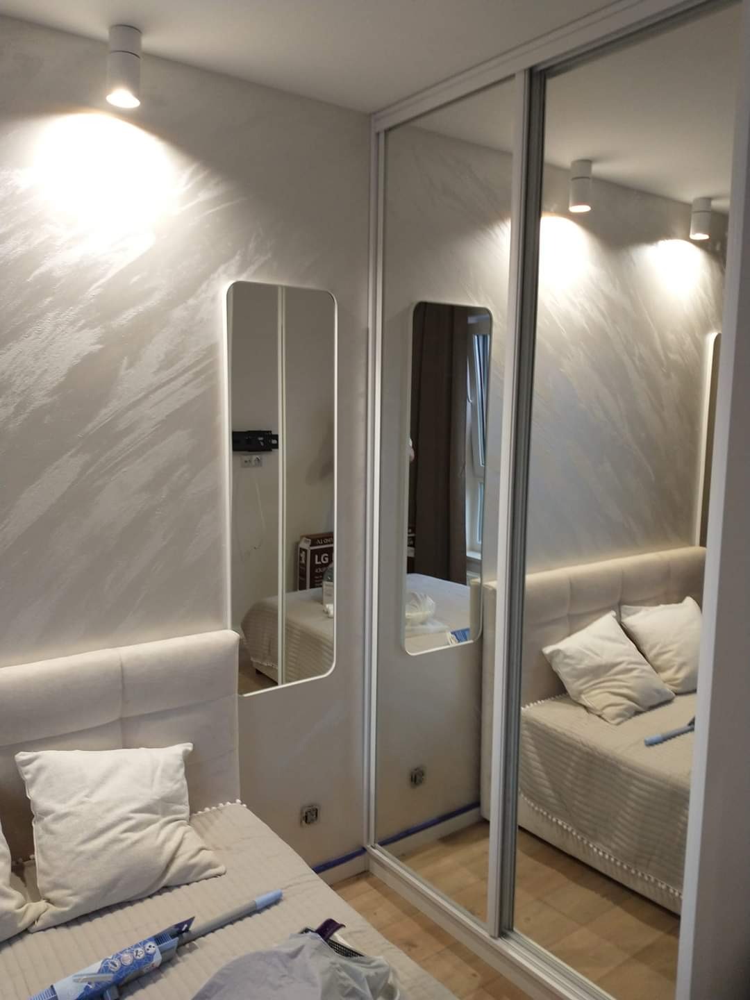 Sypialnia z łóżkiem, lustrem na ścianie i szafą przesuwną z lustrzanymi drzwiami, wzorzysta ściana w odcieniach beżu, widoczny fragment podłogi i narzędzie malarskie.